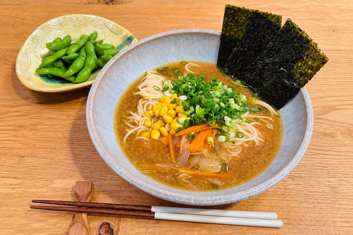 A bowl of vegan miso ramen with sweet corn, scallion, carrot and nori, beside a small dish of edamame on a wooden table